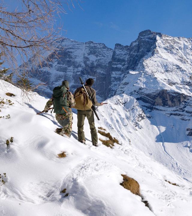 Das Bild zeigt zwei Personen, die sich in einer winterlichen Berglandschaft befinden, wahrscheinlich im Wipptal in Tirol. Die Szene ist von Schnee und steilen Berggipfeln geprägt, die im Hintergrund sichtbar sind.   Die erste Person trägt eine camouflagefarbene Jacke und hat einen großen Rucksack auf dem Rücken. Die zweite Person hat eine graue Jacke an und trägt in der Hand eine Waffe oder ein Jagdgerät. Beide stehen auf einem schneebedeckten Hang und scheinen die Landschaft zu erkunden. Der Himmel ist klar und blau, und die Sonne beleuchtet die Szenerie, wodurch Schatten auf dem Schnee entstehen.   Im Vordergrund sind einige schneebedeckte Hügel und vereinzelte grüne Pflanzen sichtbar, die aus dem Schnee ragen. Im Hintergrund befinden sich mächtige Felsen und Gipfel der Stubaier Alpen, die für diese Region charakteristisch sind.   Insgesamt vermittelt das Bild eine Atmosphäre von Abenteuer und Erkundung in der Natur.