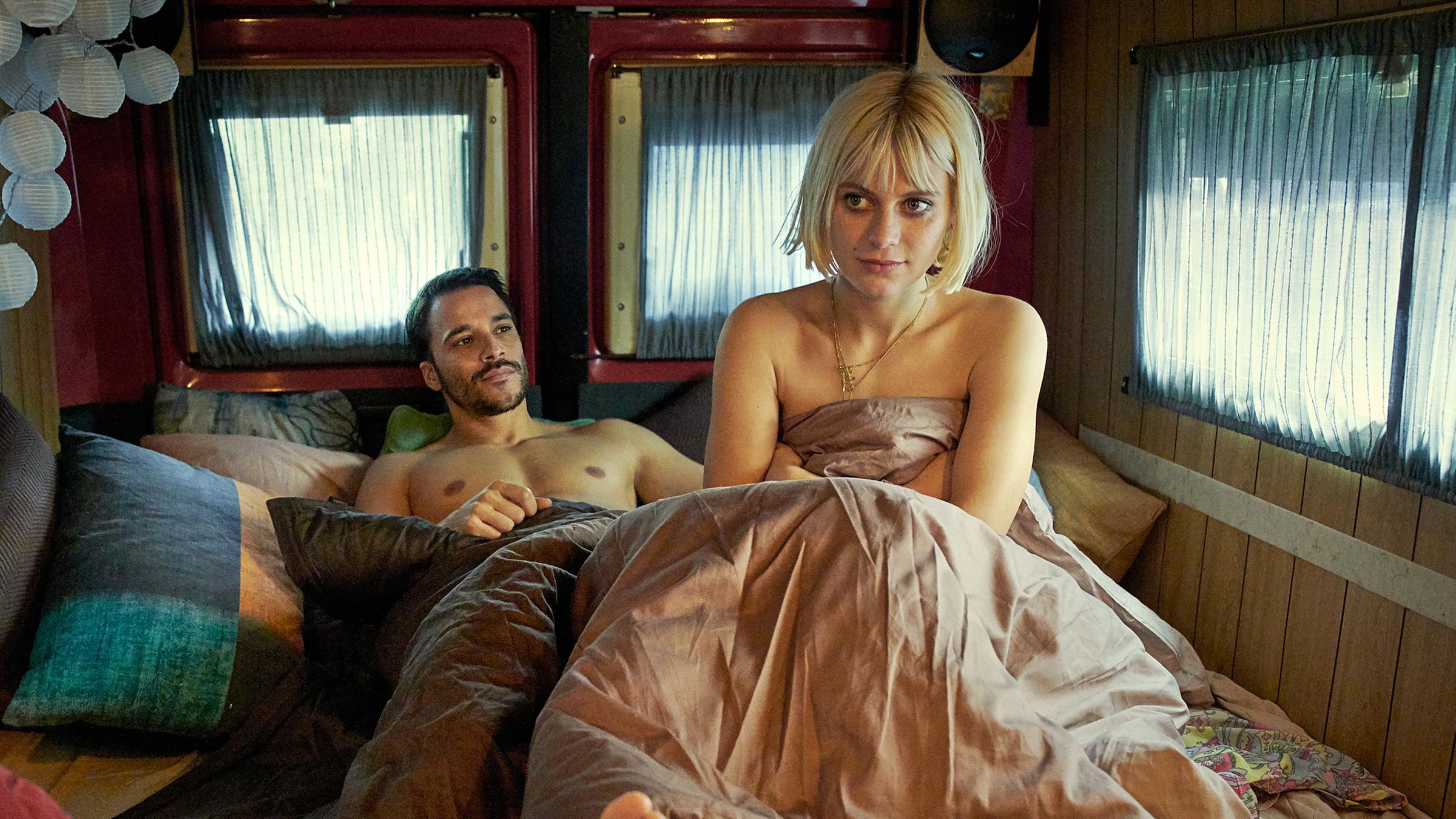 Liegender Sam (Kostja Ullmann) und sitzende Edda (Alli Neumann) nackt in Bettdecke gehüllt auf Bett im Wohnwagen.
