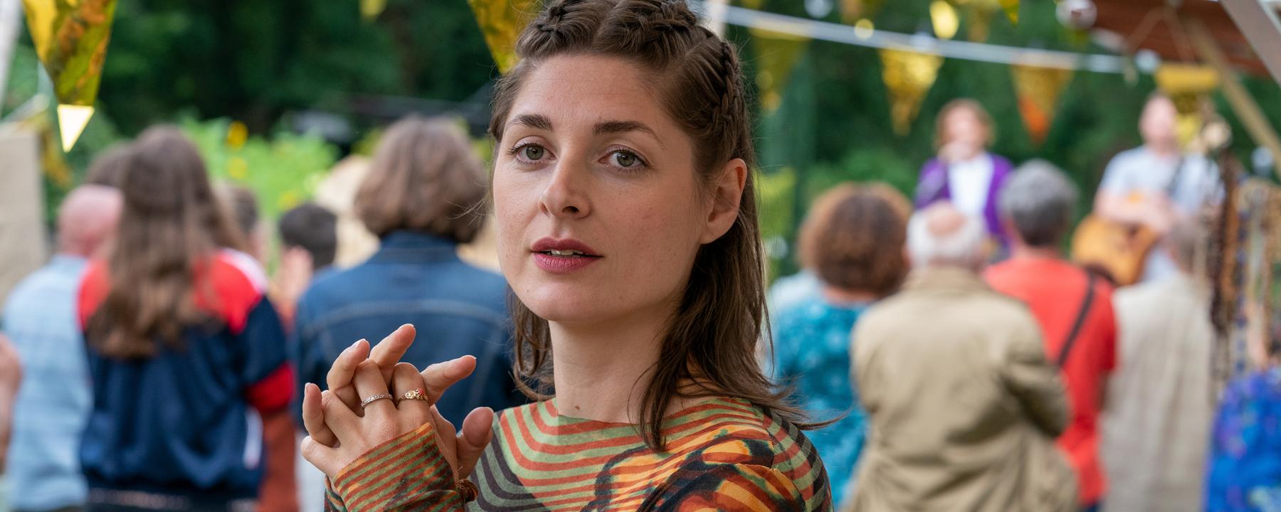 "WIR - Kürbiskönigin": Helena (Katharina Nesytowa) blickt suchend in die Ferne und hält dabei ihre Hände gefaltet vor sich. Im Hintergrund ist unscharf eine Bühne mit Musiker*innen und Zuschauer*innen zu sehen.