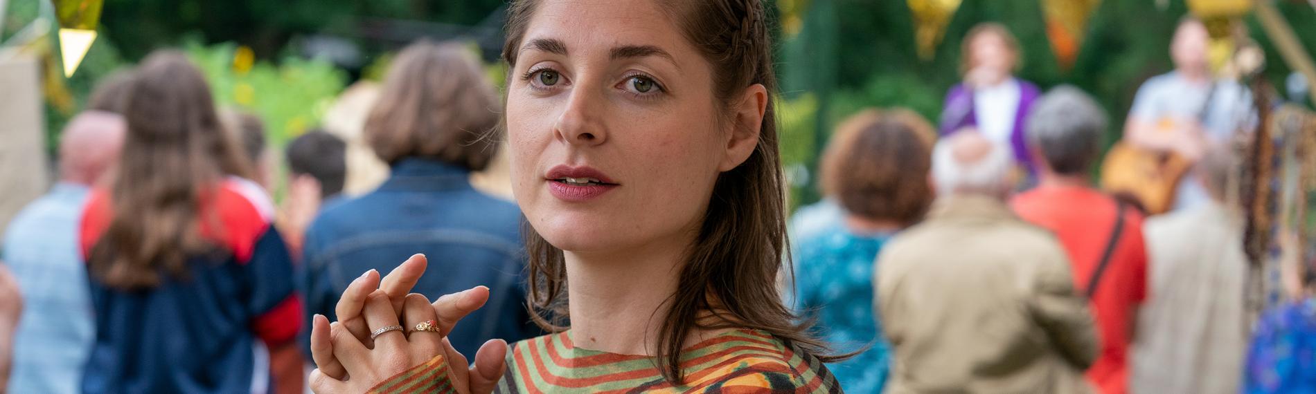 "WIR - Kürbiskönigin": Helena (Katharina Nesytowa) blickt suchend in die Ferne und hält dabei ihre Hände gefaltet vor sich. Im Hintergrund ist unscharf eine Bühne mit Musiker*innen und Zuschauer*innen zu sehen.