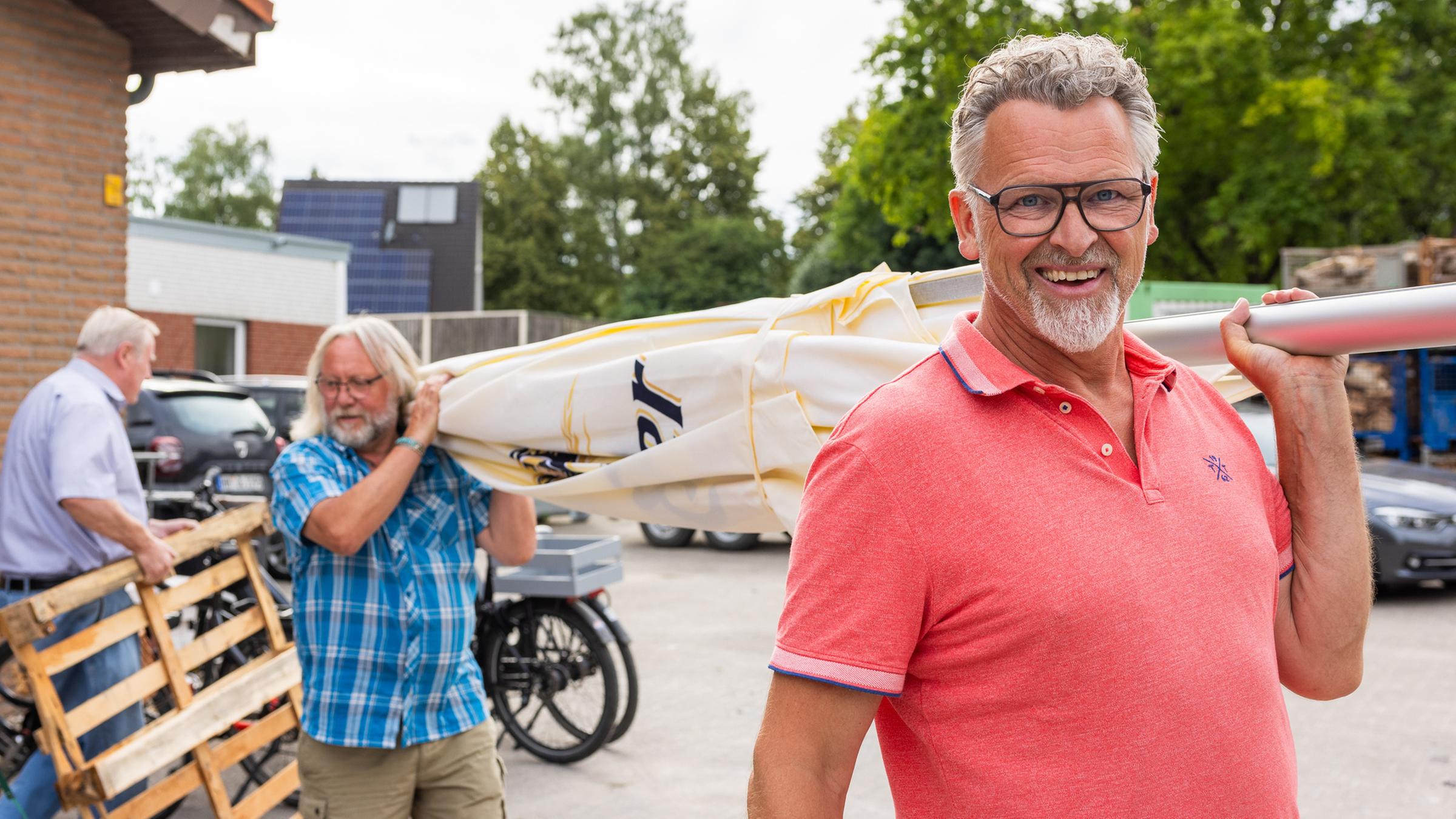 "37°: Wir retten unser Dorf - Zusammenleben auf dem Land": Zwei Männer tragen zusammen einen übergroßen Sonnenschirm auf ihren Schultern. Der im Bild vorne rechts stehende Mann trägt ein lachsfarbenes Poloshirt, eine Brille, hat dunkles graumeliertes Haar und lächelt in die Kamera.