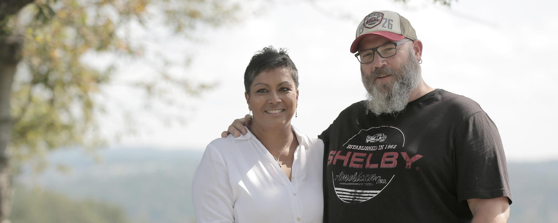 Regina und Björn Perder stehen auf einer Wiese, im Hintergrund Weitblick in die Natur. Er, ein großer, stämmiger Mann mit Vollbart, Käppi und schwarzem T-Shirt, hat seinen Arm um ihre Schulter gelegt. Sie, etwas dunklerer Teint, in weißer Bluse mit Silberarmreifen, -Halskette und -Ohrringen, legt sachte eine Hand auf seinen Bauch. Beide schauen in die Kamera.
