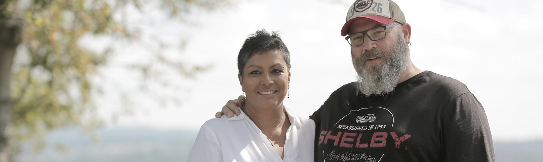 Regina und Björn Perder stehen auf einer Wiese, im Hintergrund Weitblick in die Natur. Er, ein großer, stämmiger Mann mit Vollbart, Käppi und schwarzem T-Shirt, hat seinen Arm um ihre Schulter gelegt. Sie, etwas dunklerer Teint, in weißer Bluse mit Silberarmreifen, -Halskette und -Ohrringen, legt sachte eine Hand auf seinen Bauch. Beide schauen in die Kamera.