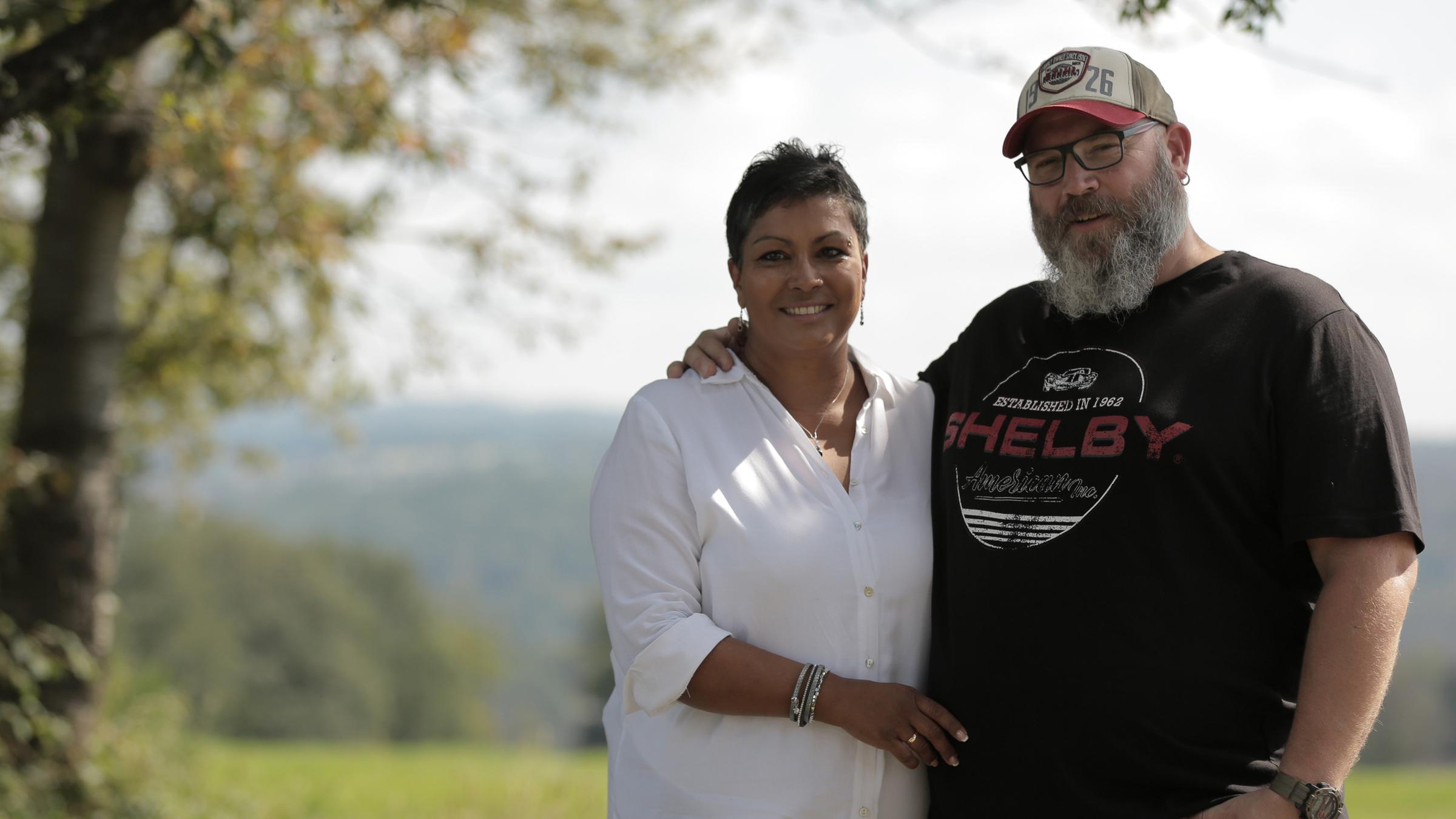Regina und Björn Perder stehen auf einer Wiese, im Hintergrund Weitblick in die Natur. Er, ein großer, stämmiger Mann mit Vollbart, Käppi und schwarzem T-Shirt, hat seinen Arm um ihre Schulter gelegt. Sie, etwas dunklerer Teint, in weißer Bluse mit Silberarmreifen, -Halskette und -Ohrringen, legt sachte eine Hand auf seinen Bauch. Beide schauen in die Kamera.