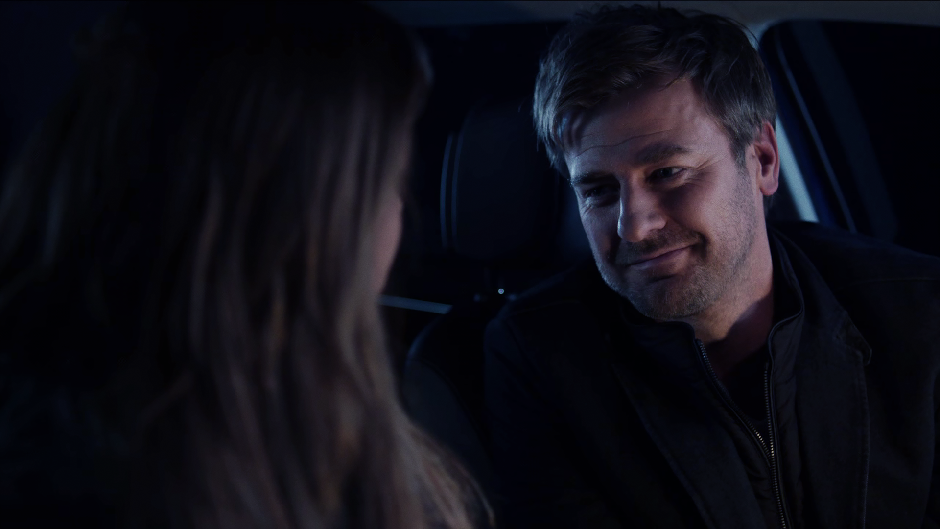 Harald Neuhauser (Marcus Mittermeier) sitzt in Auto und lächelt eine Frau an, die nur von hinten zu sehen ist. 