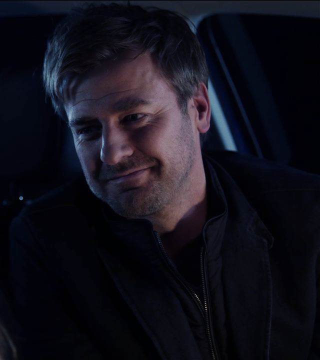 Harald Neuhauser (Marcus Mittermeier) sitzt in Auto und lächelt eine Frau an, die nur von hinten zu sehen ist. 