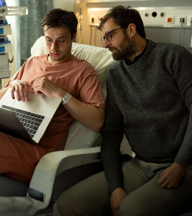 "The Kollective - Wir sind Millionen": Lucas (Gijs Blom) sitzt aufrecht im Krankenbett und blickt auf seinen aufgeklappten Laptop. Neben ihm sitzt Etienne (Grégory Montel) und schaut ebenfalls in die Richtung des Computers.