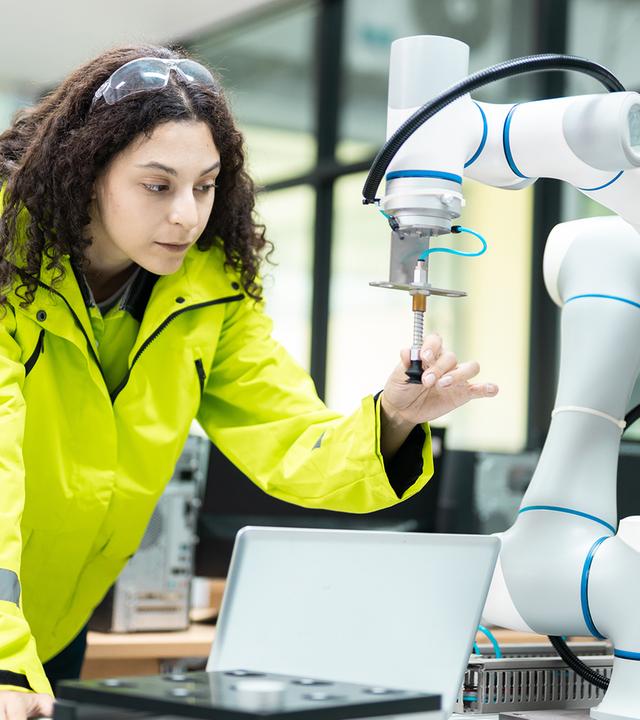 Eine Frau in einer Werkhalle oder einem Labor beugt sich über einen Tisch und arbeitet konzentriert an einem weißen Robotarm mit Greifwerkzeug. Sie trägt eine auffällige gelbe Schutzjacke und blickt auf ein Bauteil, das der Roboter hält. Neben ihr steht ein Laptop, im Hintergrund sind technische Geräte zu sehen. Das Bild zeigt die Entwicklung und Anwendung moderner Robotik.