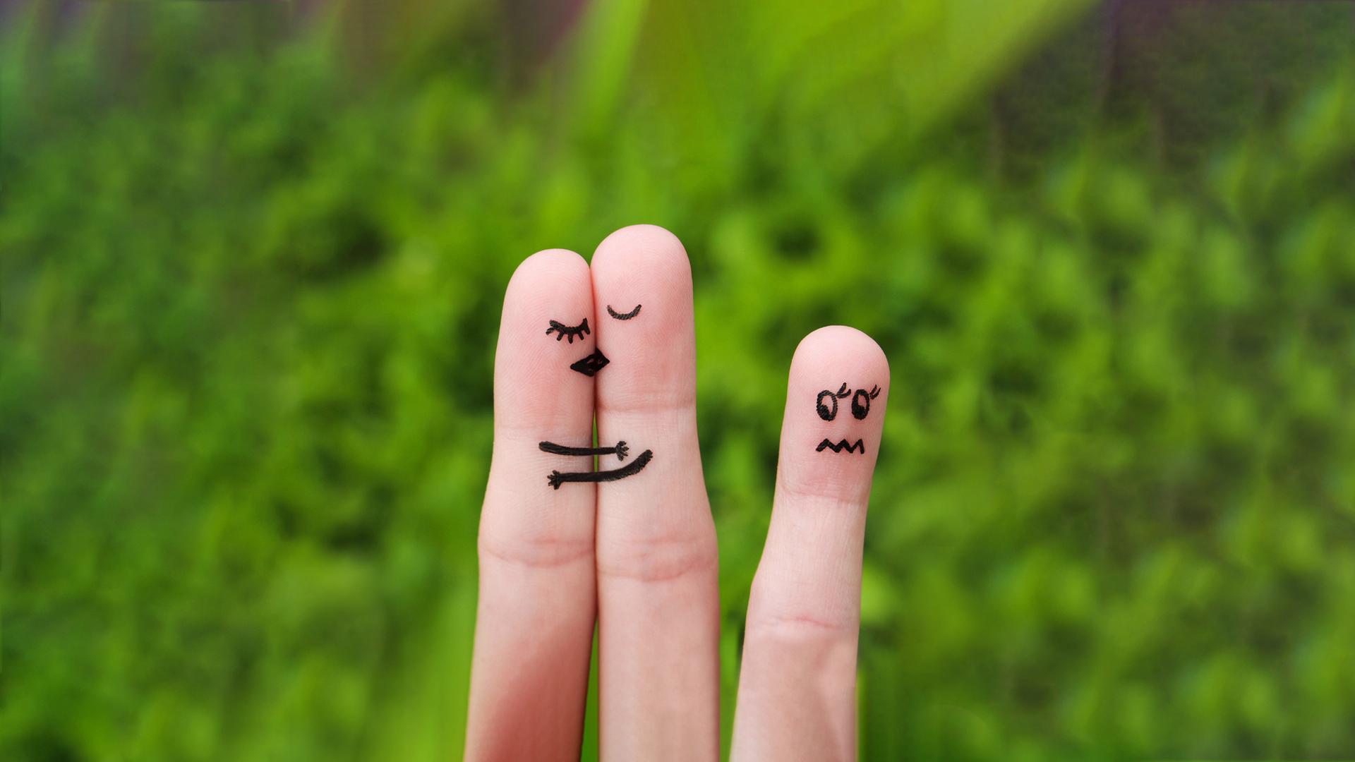 zwei Finger schmiegen sich aneinander, auf Ihnen die Gesichtsmimik von sich Küssenden aufgemalt, ein einzelner Finger mit aufgemalter unglücklicher Mimik im Abseits