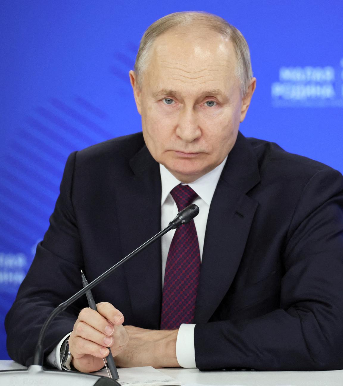 Wladimir Putin bei einem Treffen mit Gemeindevorstehern. Putin blickt in die Kamera. Er hält einen Kugelschreiber in der Hand. Hintergrund ist blau.