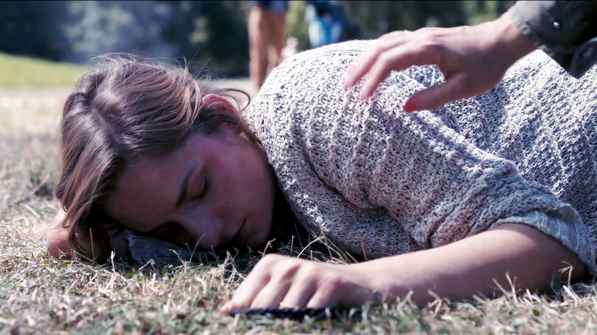 Angelika Flierl (Bernadette Heerwagen) liegt bäuchlings auf einem Stück Rasen, scheinbar bewusstlos. Eine Hand greift nach ihrer Schulter. 