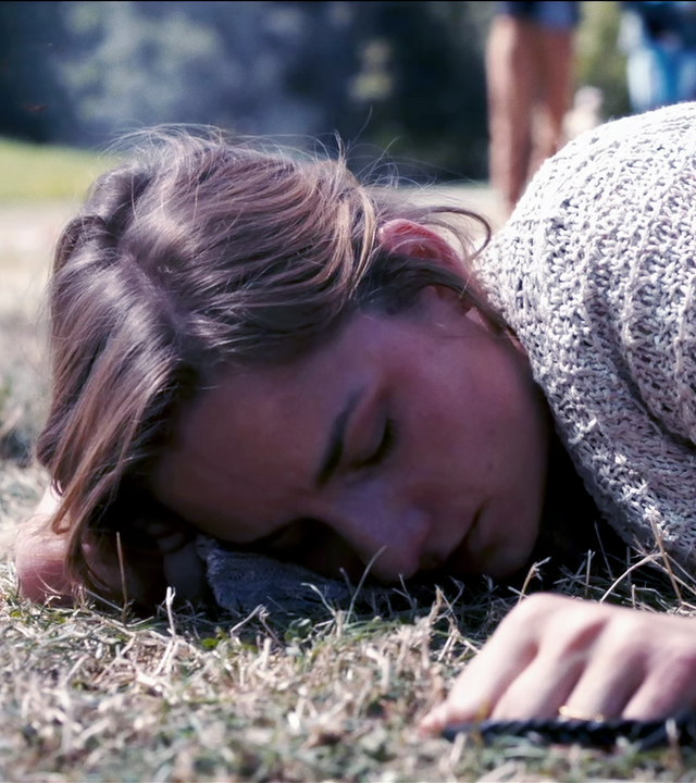 Angelika Flierl (Bernadette Heerwagen) liegt bäuchlings auf einem Stück Rasen, scheinbar bewusstlos. Eine Hand greift nach ihrer Schulter. 