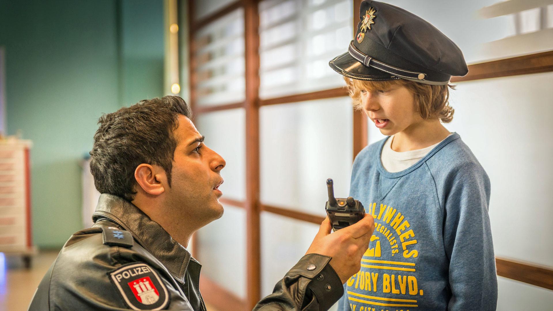 "Notruf Hafenkante - Wo ist Papa?": Auf dem Flur des Elbkrankenhauses kniet der Polizist Tarik Coban (Serhat Cokgezen) vor einem kleinen Jungen (Arsseni Bultmann), der eine Polizeimütze trägt. Tarik hält dem Kleinen das Funkgerät hin.