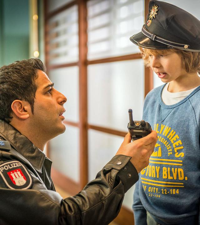 "Notruf Hafenkante - Wo ist Papa?": Auf dem Flur des Elbkrankenhauses kniet der Polizist Tarik Coban (Serhat Cokgezen) vor einem kleinen Jungen (Arsseni Bultmann), der eine Polizeimütze trägt. Tarik hält dem Kleinen das Funkgerät hin.