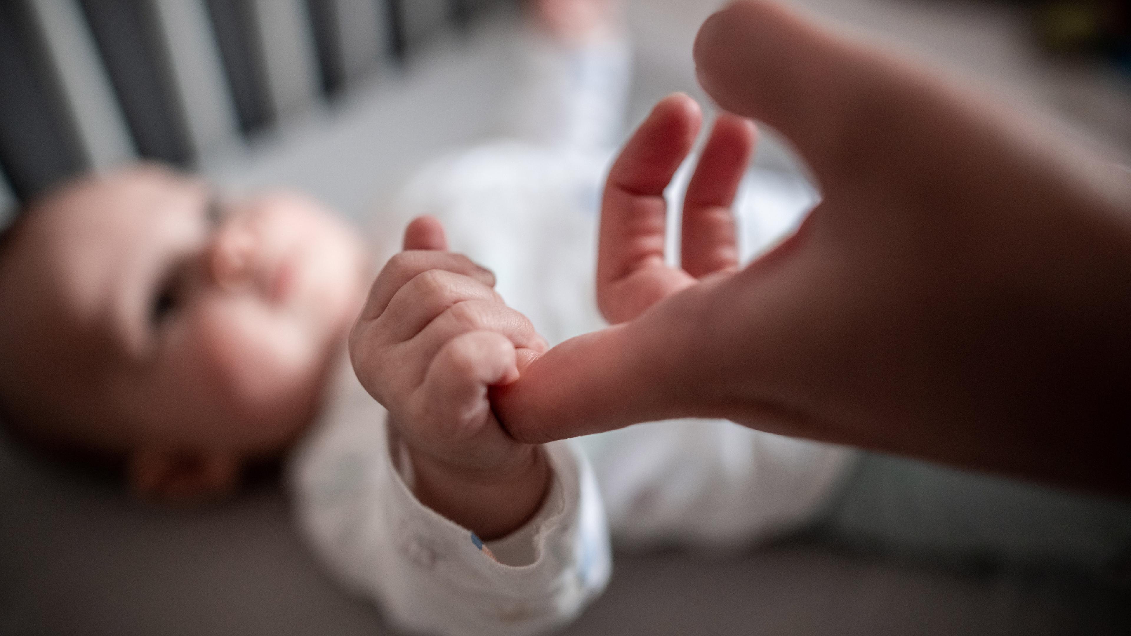 Ein Baby liegt in einem Bettchen und krallt sich am Zeigefinger der Mutter fest.
