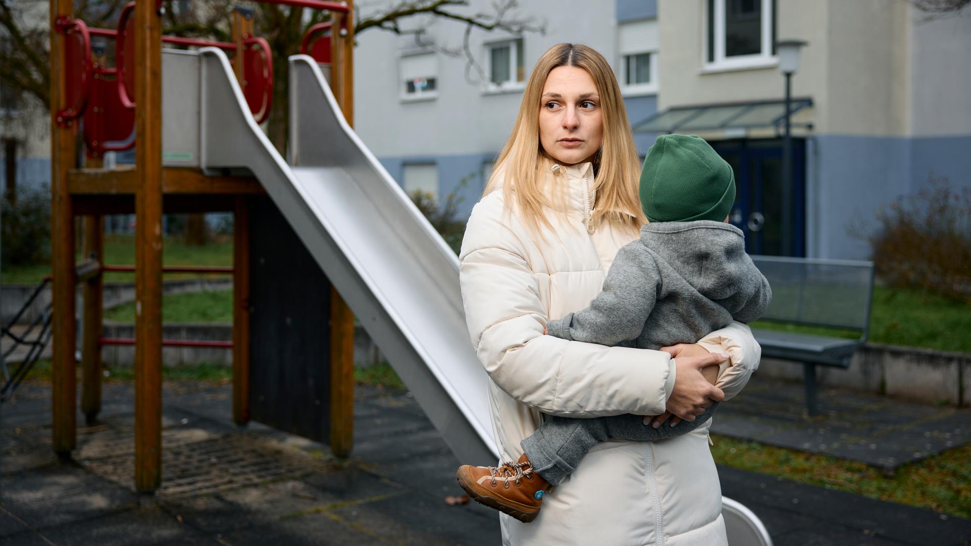 Annie hält ihr Baby auf dem Arm und schaut traurig und erschöpft zur Seite. Im Hintergrund ist ein Spielplatz mit einer Rutsche zu sehen.