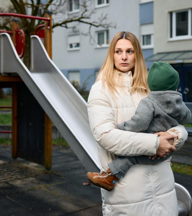 Annie hält ihr Baby auf dem Arm und schaut traurig und erschöpft zur Seite. Im Hintergrund ist ein Spielplatz mit einer Rutsche zu sehen.