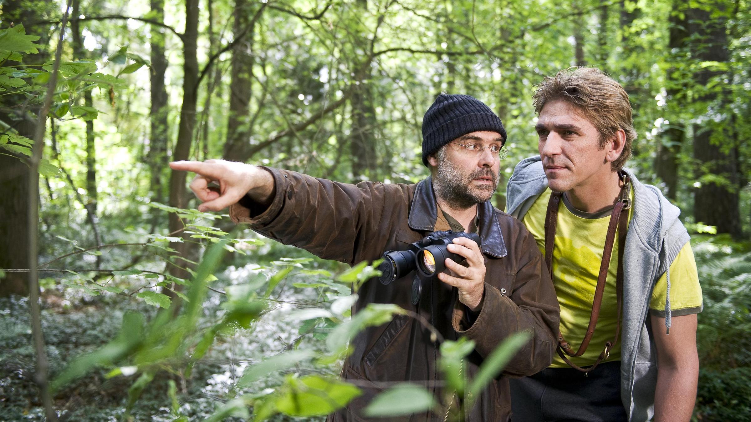 Wolfsforscher Janosch Blumkov (Jophi Ries, links) und Fritz Fuchs (Guido Hammesfahr, rechts) stehen zusammen mit Keks im Wald. Blumkov zeigt mit ausgestrecktem Arm nach links.