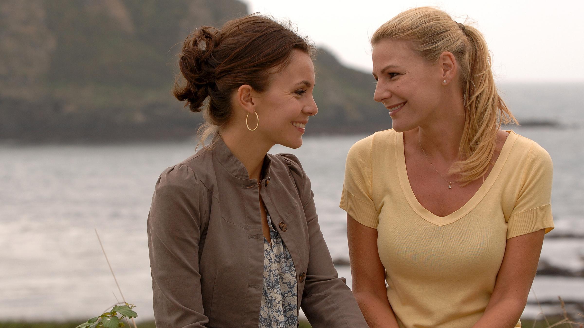 Sabrina (Nadine Warmuth) und Lisa (Sophie Schütt) sitzen am Ufer nebeneinander und lächeln sich an.