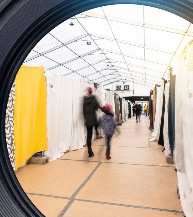Blick in den Gang eines Flüchtlingszeltes. In der Mitte verschwommen zwei Personen. Links und rechts hängen Vorhänge als Blickschutz.