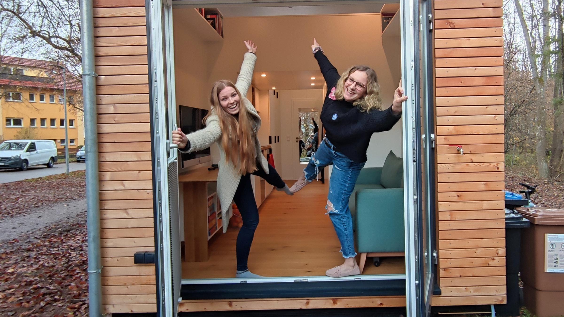 Reporterin Wiebke Denker und Joey stehen an der Tür eines Tiny-House. Sie strecken jede ein Bein und einen Arm in die Luft. Mit den anderen Armen halten sie sich an den geöffneten Türen fest. Beide lachen.