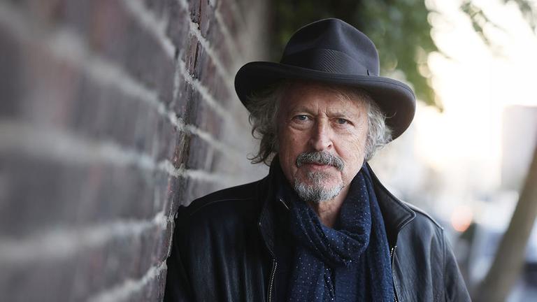 Mann mit Hut steht an einer Mauer und schaut in die Kamera