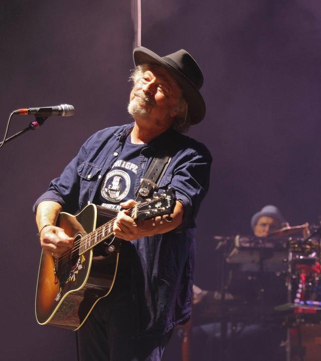 Ein älterer Mann mit Bart und Hut spielt Gitarre auf einer Bühne und lächelt.