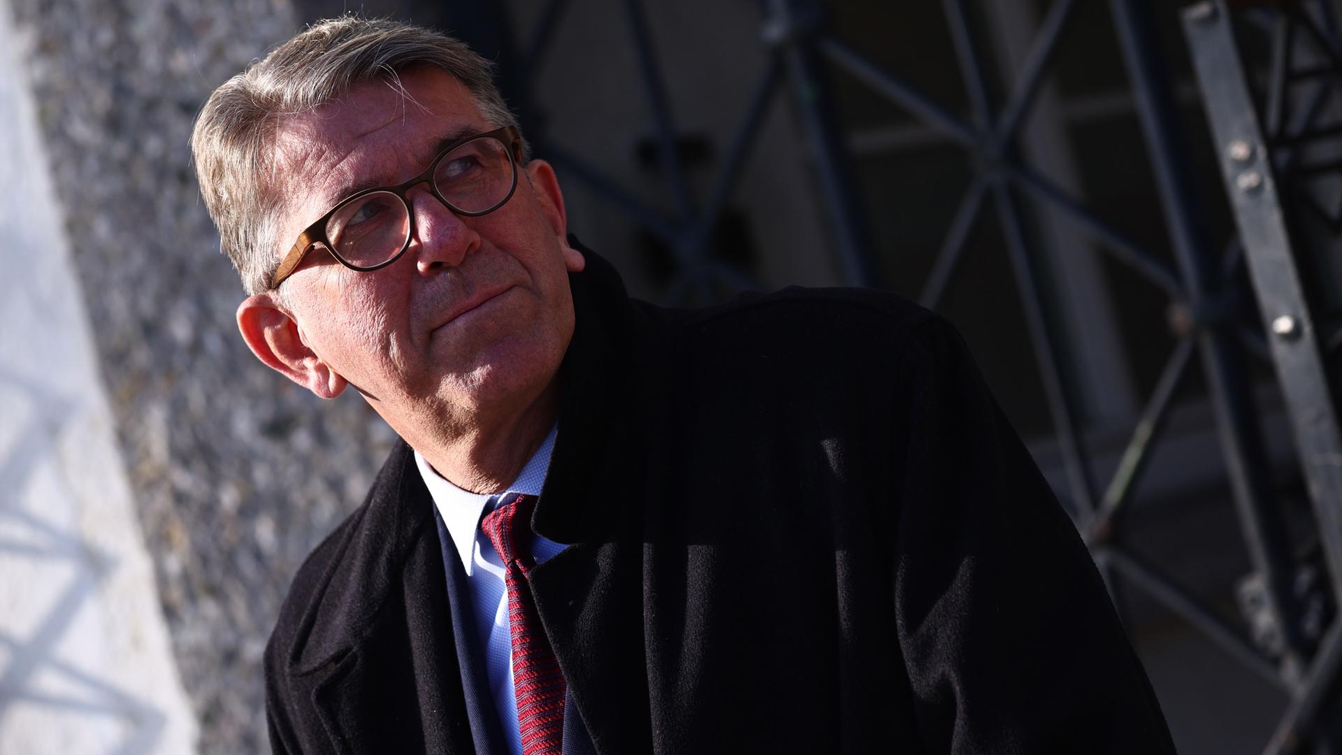 Ein Mann mit graumelierten kurzen Haaren, Brille und im dunklen Anzug und Mantel schaut nach links.