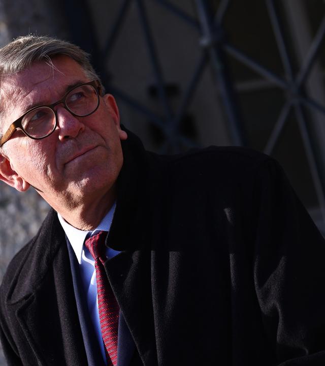 Ein Mann mit graumelierten kurzen Haaren, Brille und im dunklen Anzug und Mantel schaut nach links.