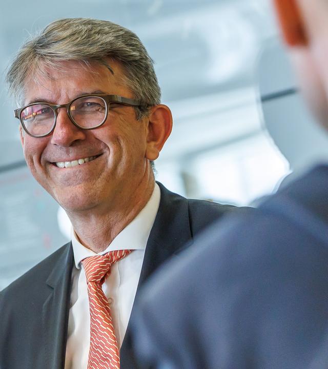 Foto eines Mannes im Anzug mit runder Brille und kurzen graumelierten blonden Haaren, der lächelt.