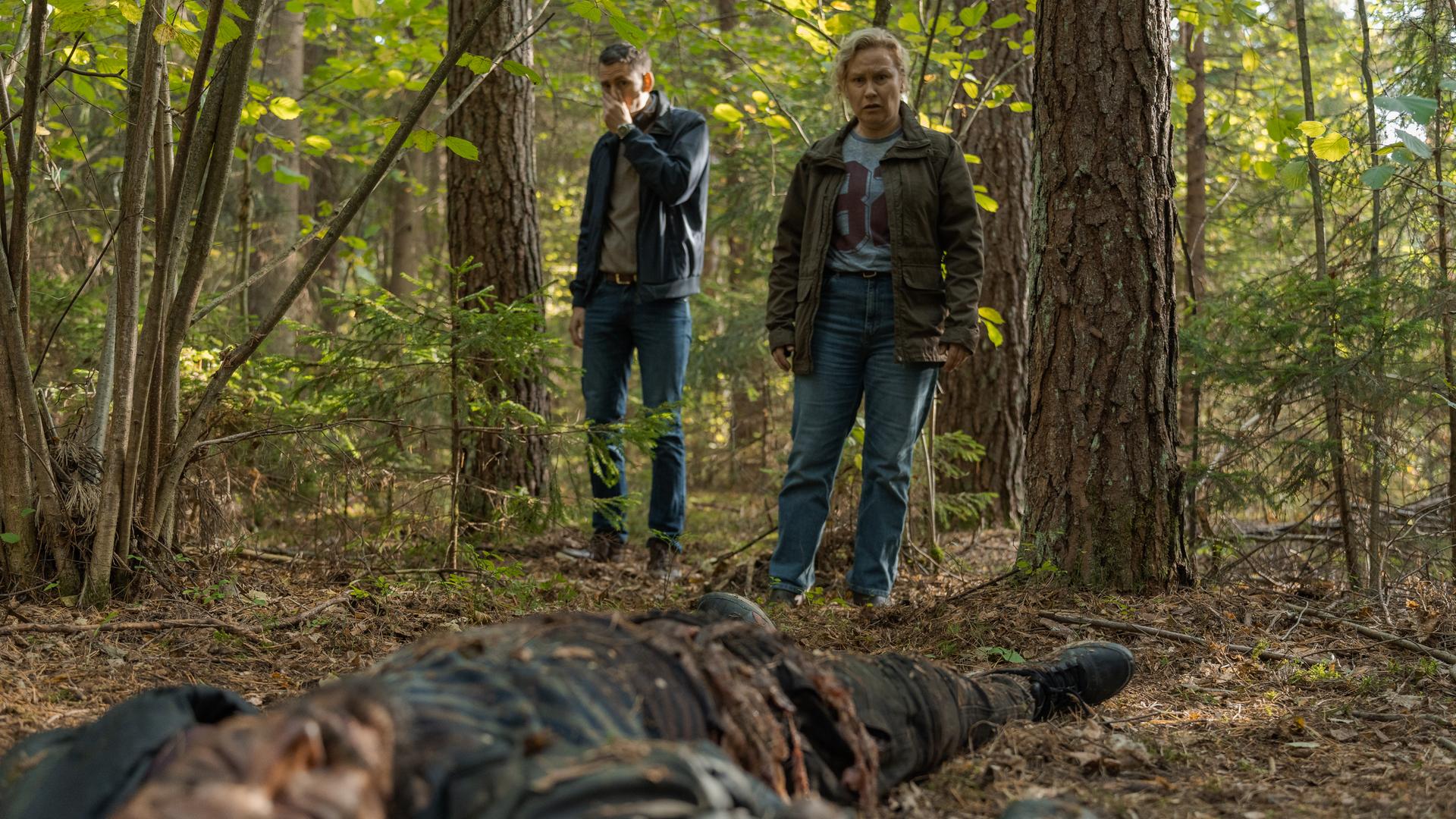 "Wolfssommer - Blutige Spuren: Die Spur der Wölfe": Die Leiche von Vadim (Mykolas Vildžiūnas) liegt im Wald auf dem Boden. Hannah (Eva Melander) und Gordon (Hannes Fohlin) stehen zwischen zwei Bäumen und betrachten den Toten. Gordon hält sich die linke Hand vor Mund und Nase.