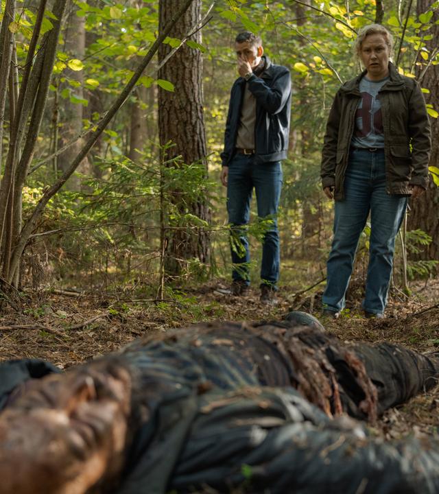 "Wolfssommer - Blutige Spuren: Die Spur der Wölfe": Die Leiche von Vadim (Mykolas Vildžiūnas) liegt im Wald auf dem Boden. Hannah (Eva Melander) und Gordon (Hannes Fohlin) stehen zwischen zwei Bäumen und betrachten den Toten. Gordon hält sich die linke Hand vor Mund und Nase.