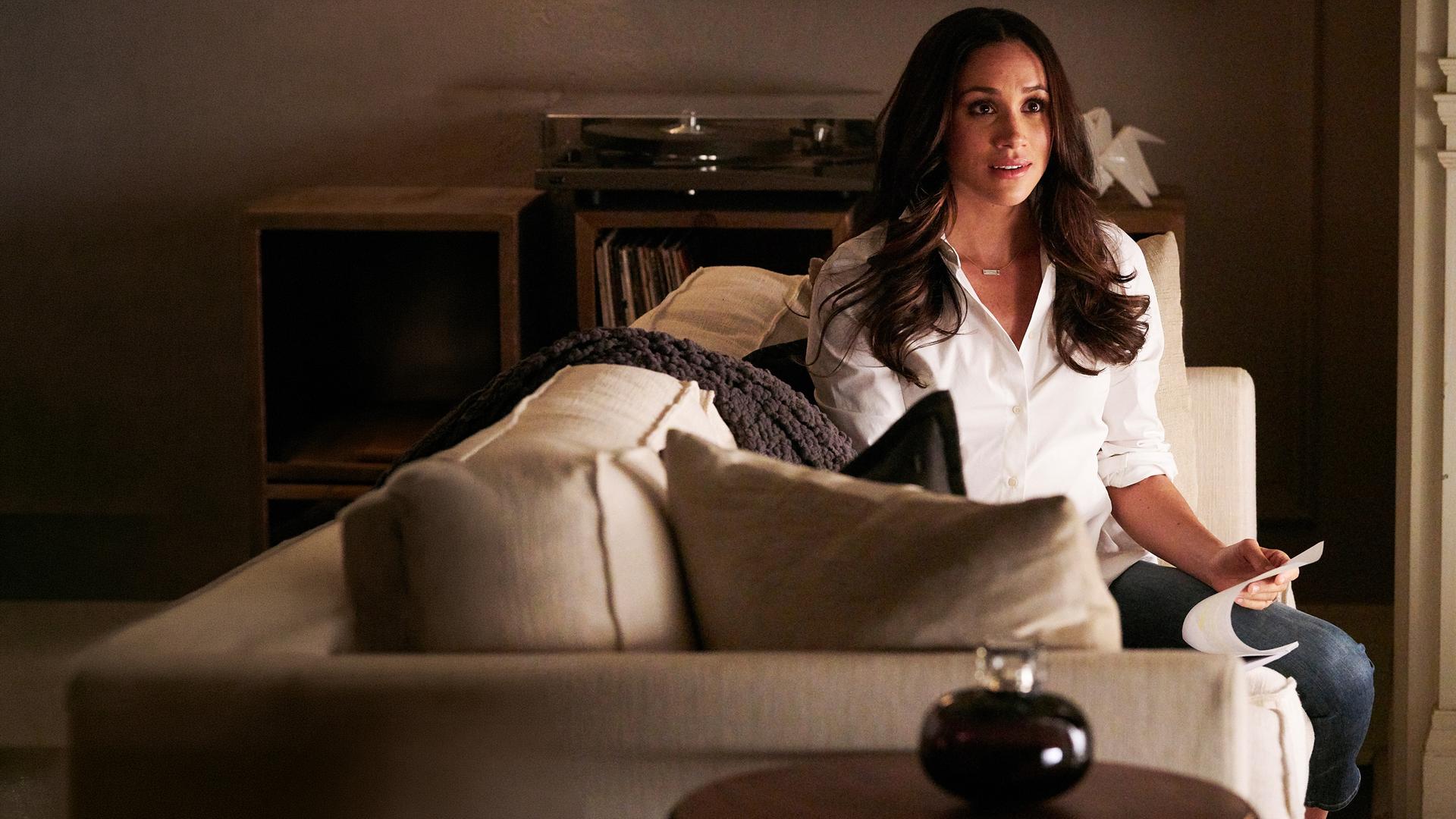 "Suits - Wortbruch": Rachel (Meghan Markle) sitzt auf einem cremefarbenen Sofa mit einer weißen Bluse bekleidet. Sie blättert in einem Dokument und schaut dabei erwartungsvoll auf.