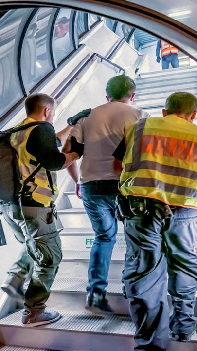 Montage: Ein Mann wird von zwei Polizisten eine Gangway zu einem Flugzeug hochgeführt. Dahinter ist im Cut-out-Stil ein schwarzer Rand mit Papierkante.