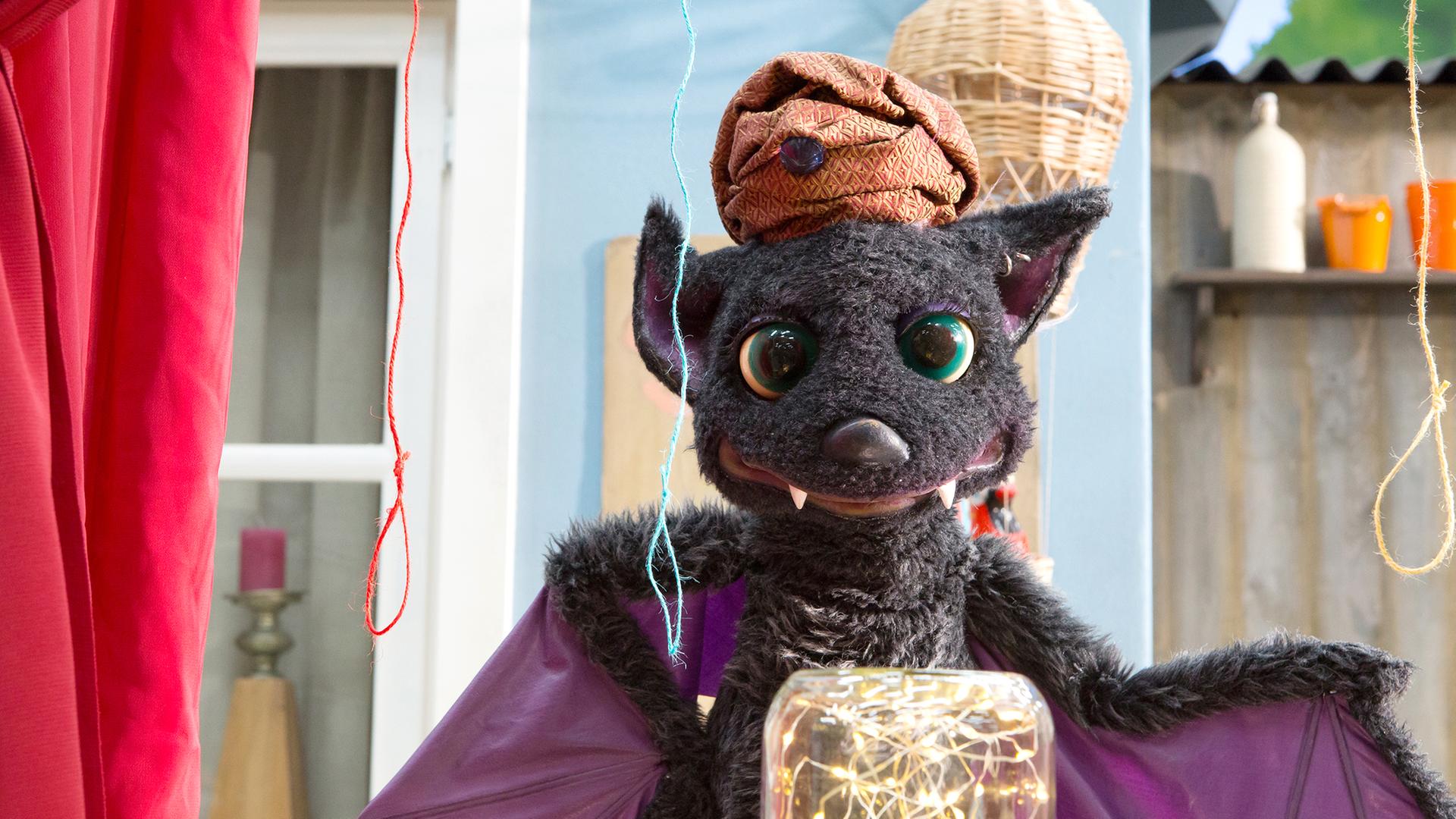 Vampi hat einen Turban auf dem Kopf und steht beschwörend vor einem Einmachglas, in dem ein Lichterkettchen funkelt. Über ihr hängen drei Schnüren in unterschiedlichen Farben.