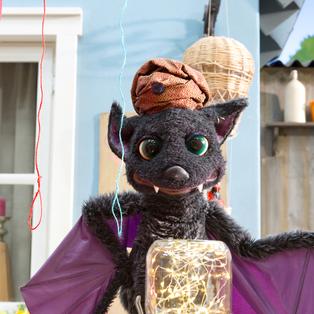 Vampi hat einen Turban auf dem Kopf und steht beschwörend vor einem Einmachglas, in dem ein Lichterkettchen funkelt. Über ihr hängen drei Schnüren in unterschiedlichen Farben.