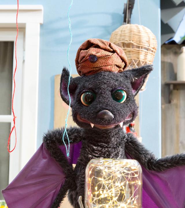 Vampi hat einen Turban auf dem Kopf und steht beschwörend vor einem Einmachglas, in dem ein Lichterkettchen funkelt. Über ihr hängen drei Schnüren in unterschiedlichen Farben.