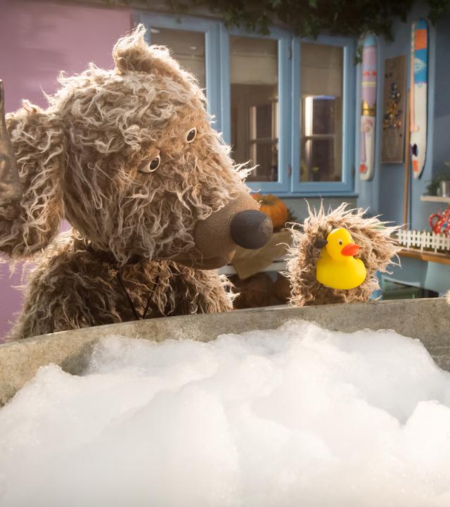 Hund Buffo steht vor der Badewanne und hat eine Quietscheente in der Hand. Wurm Lotti sitzt in der Badewanne und badet.