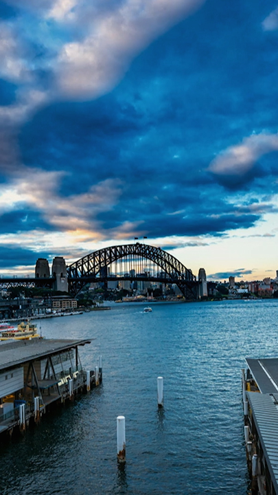 "Wunder der Baukunst (3/3) - Brücken": Die obere Bildhälfte nehmen dunkle bläuliche Wolken ein. Im Vordergrund Hafenanlagen, Kais und kleine Fährschiffe, am rechten Bildrand ein zehn stöckiges Gebäude. In der Mitte des Bildes, aber relativ unauffällig, eine große Brücke, die eine Hafeneinfahrt überspannt. Auffällig die bogenförmige Stahlkonstruktion oberhalb der Fahrbahn.