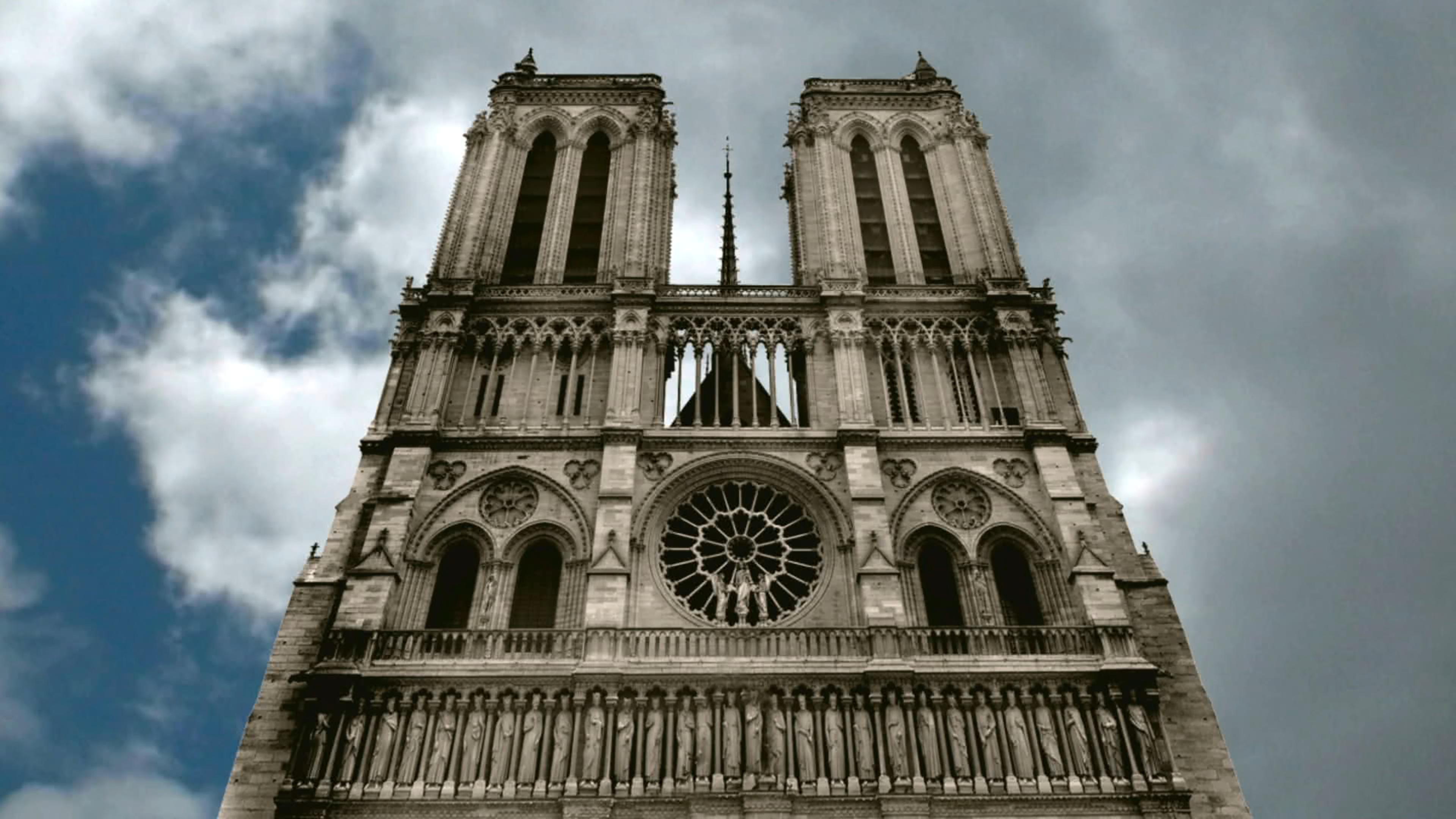 Froschperspektive auf die Front der Kathedrale von Notre-Dame, dahinter stark bewölkter Himmel.
