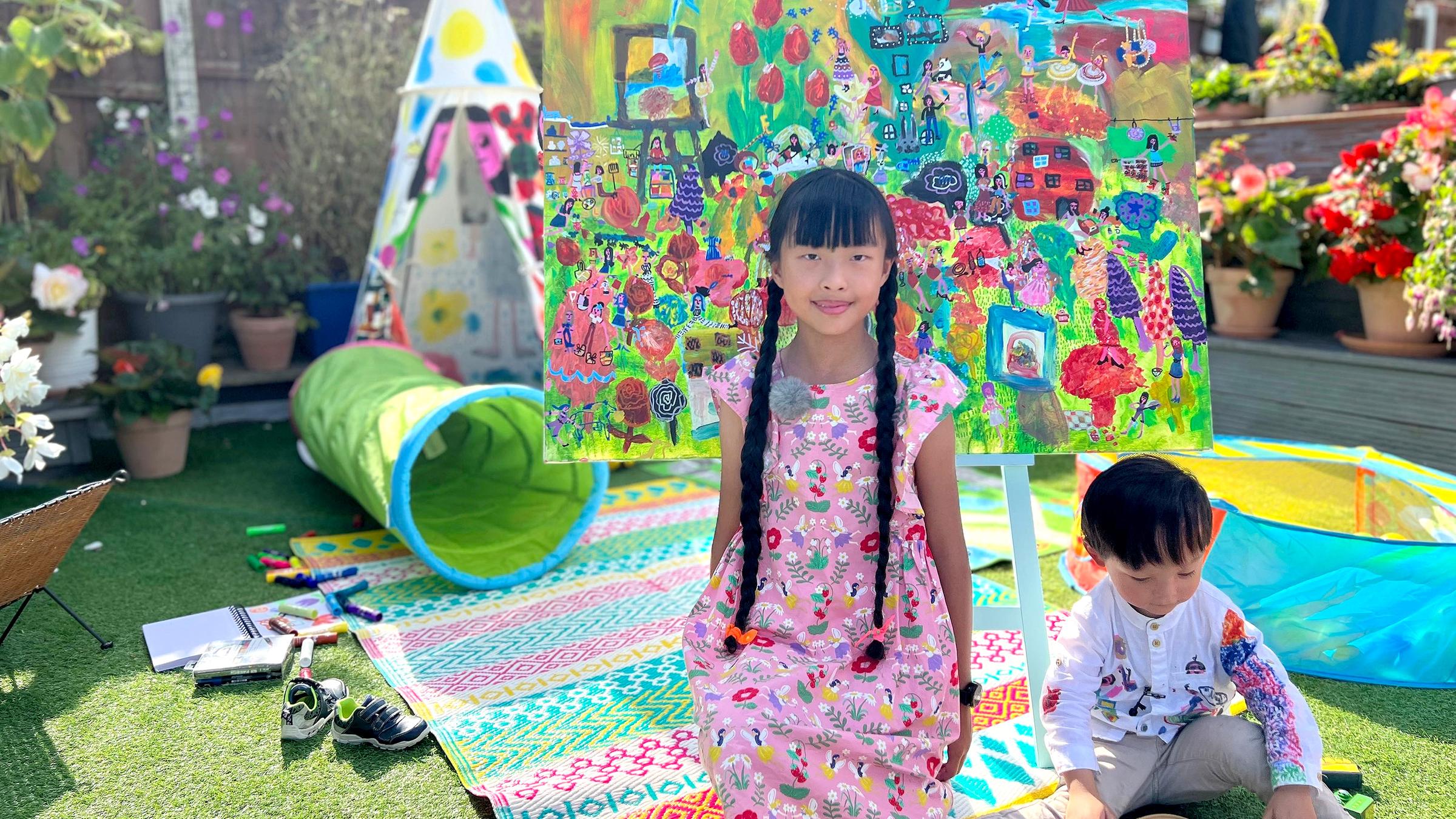 Ein Mädchen mit langen braunen Zöpfen steht vor einem großen bunten Gemälde, neben ihr ein kleiner Junge, der spielt.