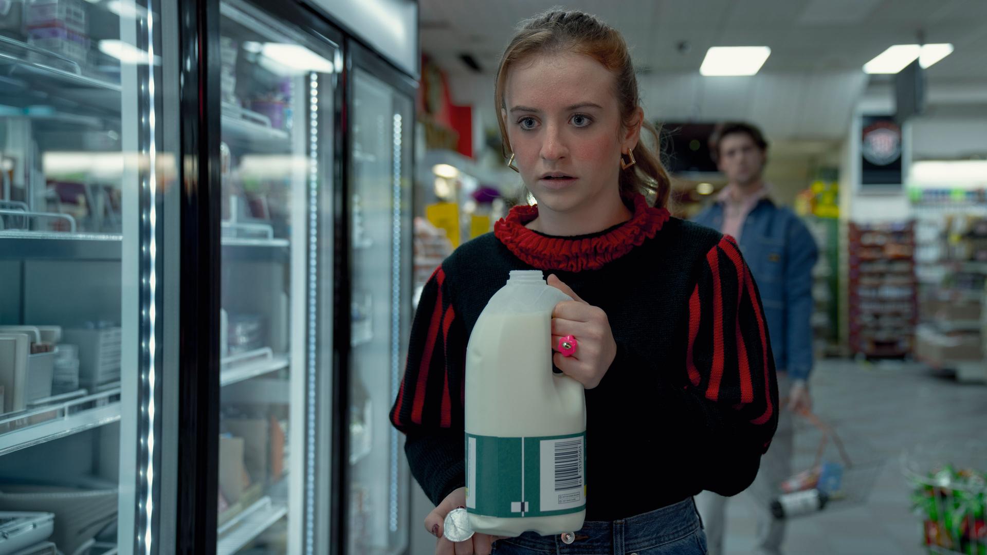 "Extraordinary - Wundermittel": Jen (Máiréad Tyers) steht mit einer geöffneten Milchflasche in den Händen in einem Supermarkt, während ihr Blick angespannt und leicht erschrocken ist. Hinter ihr steht Luke (Ned Porteous), der sie aus einiger Entfernung beobachtet und einen Einkaufskorb in der Hand hält.
