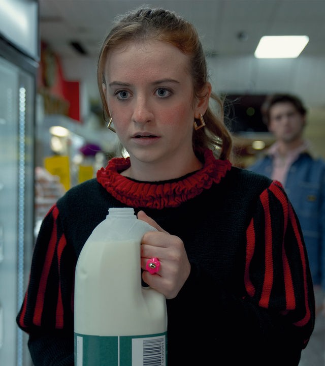 "Extraordinary - Wundermittel": Jen (Máiréad Tyers) steht mit einer geöffneten Milchflasche in den Händen in einem Supermarkt, während ihr Blick angespannt und leicht erschrocken ist. Hinter ihr steht Luke (Ned Porteous), der sie aus einiger Entfernung beobachtet und einen Einkaufskorb in der Hand hält.