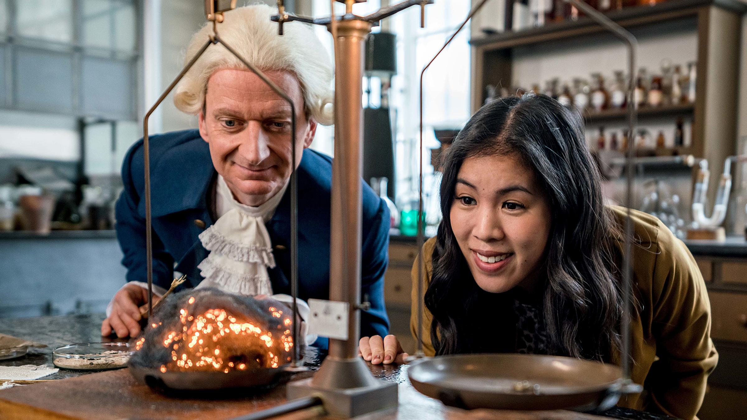Mai Thi Nguyen-Kim und Michael Kessler (als Darsteller des Wissenschaftlers Joseph Priestley) blicken in einem Labor auf eine Waage.