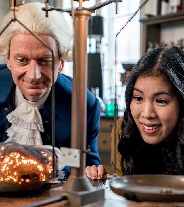 Mai Thi Nguyen-Kim und Michael Kessler (als Darsteller des Wissenschaftlers Joseph Priestley) blicken in einem Labor auf eine Waage.