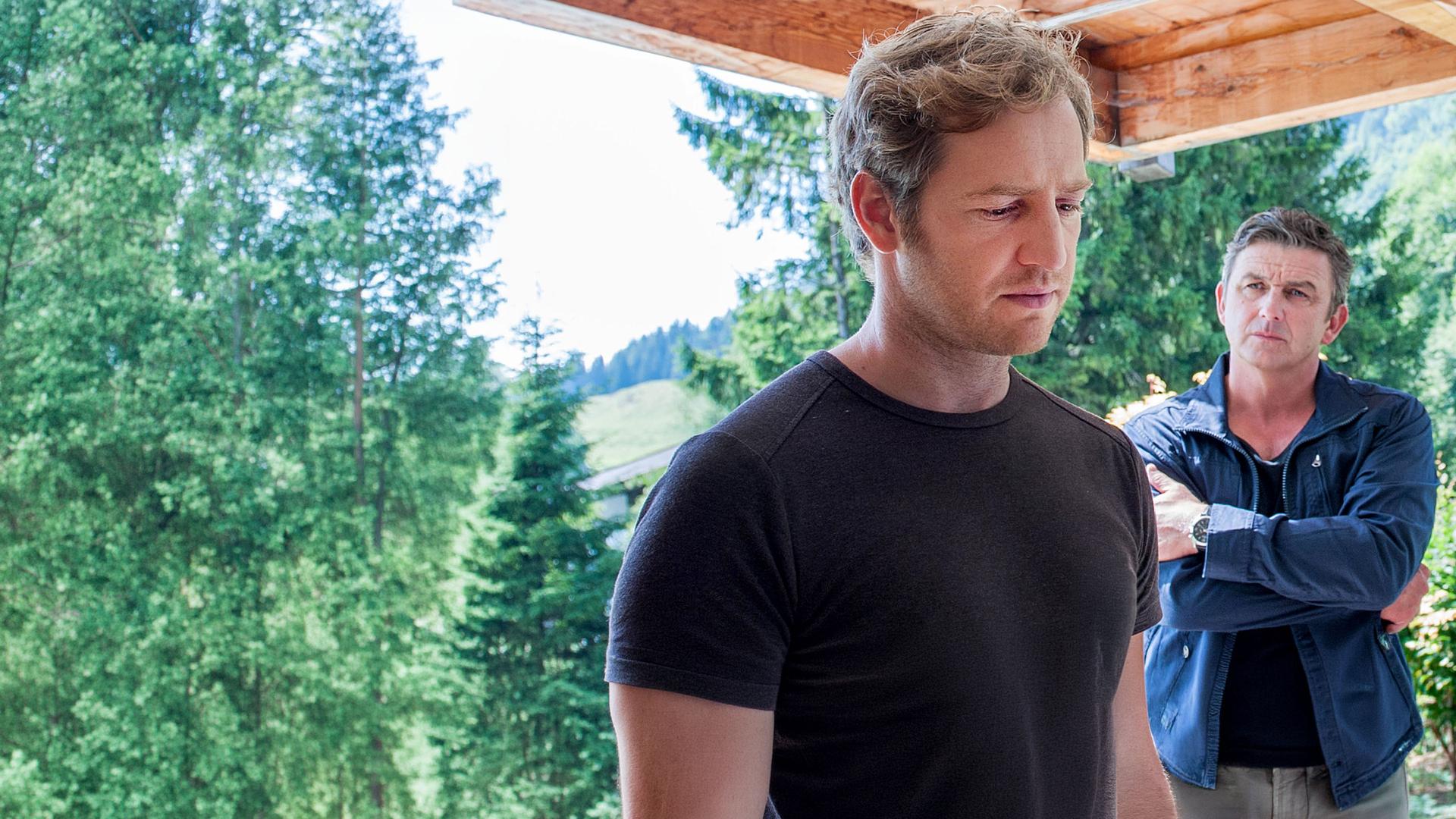 Carsten Maiwald (Tom Radisch) liest einen Brief, den er in der Hand hält. Dr. Martin Gruber (Hans Sigl) steht neben ihm auf der Terrasse und beobachtet ihn.