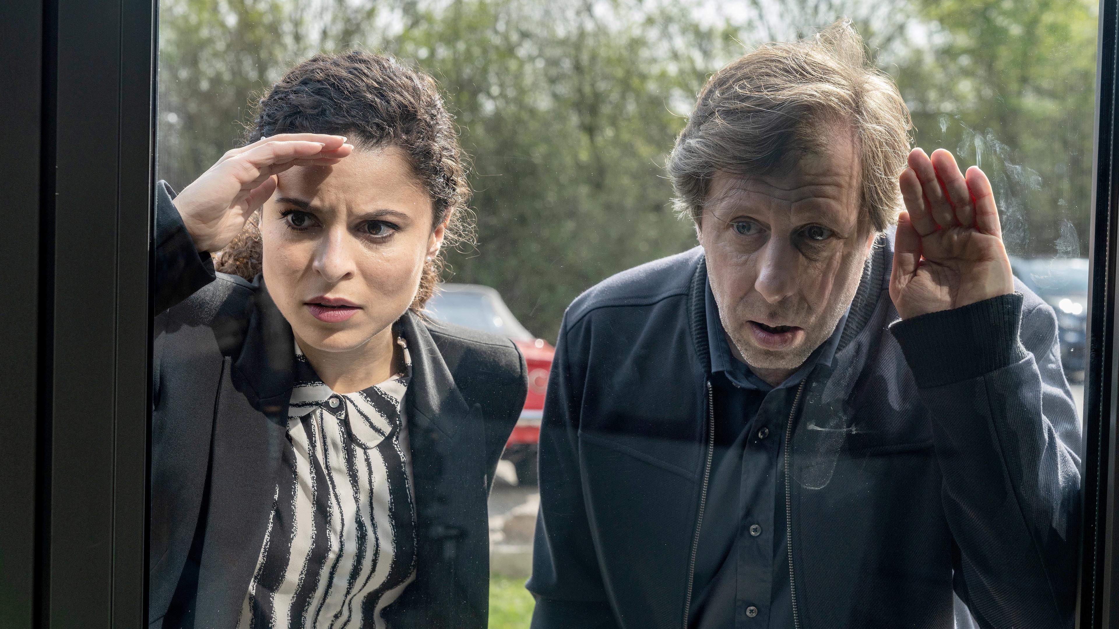 Tessa Tilker (Patricia Meeden) und Ekki (Oliver Korittke) stehen nebeneinander an einer großen Glasscheibe und schauen ans Fenster gelehnt ins Innere eines Gebäudes.