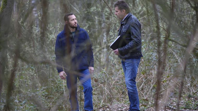 Sven Voss und Ermittler Gerhard Hoppmann stehen im Wald. Sven Voss schaut betroffen zu Ermittler Gerhard Hoppmann, während dieser ihm etwas erzählt. 