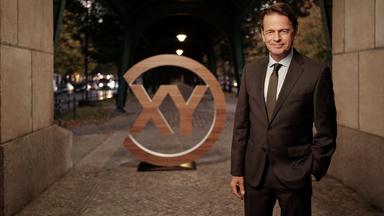 "Aktenzeichen XY... Ungelöst: Moderator Rudi Cerne":  Rudi Cerne steht vor dem "XY"-Logo und lächelt in die Kamera.