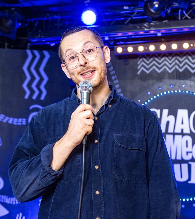 Yannick de la Pêche steht in der Mitte des Bildes. In seiner rechten Hand hält er ein Mikrofon. Er trägt eine Brille.